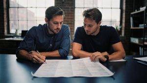 A finance lead and engineer reviewing an itemized Magento replatform cost list in a Brooklyn office
