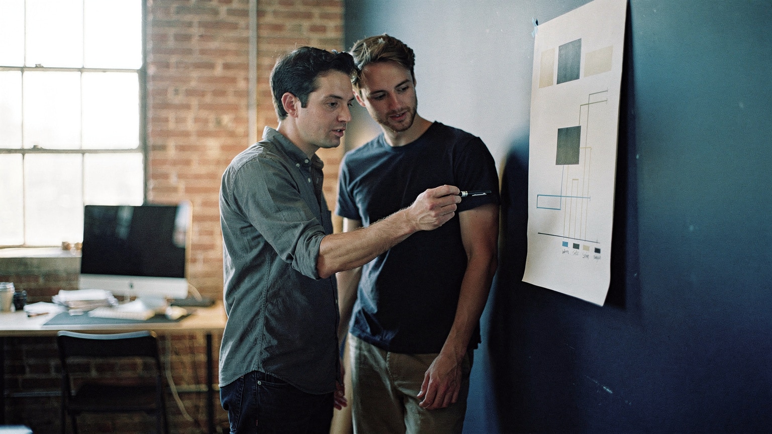A project manager reviewing a Magento project burndown on a wall chart with a developer in a Brooklyn studio