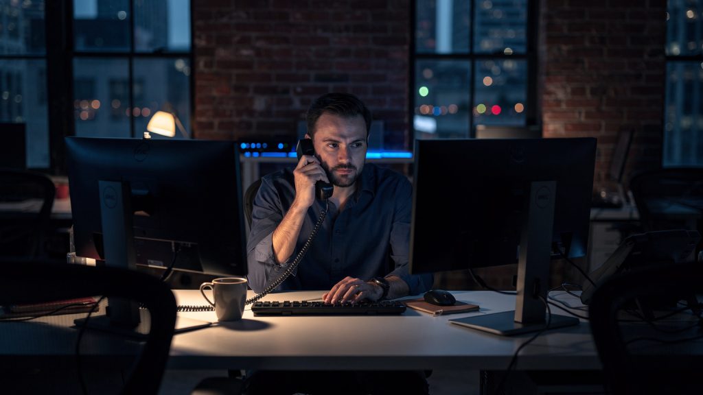 eCommerce operations director alone in a dark Brooklyn office at night on a landline phone beside powered-off monitors
